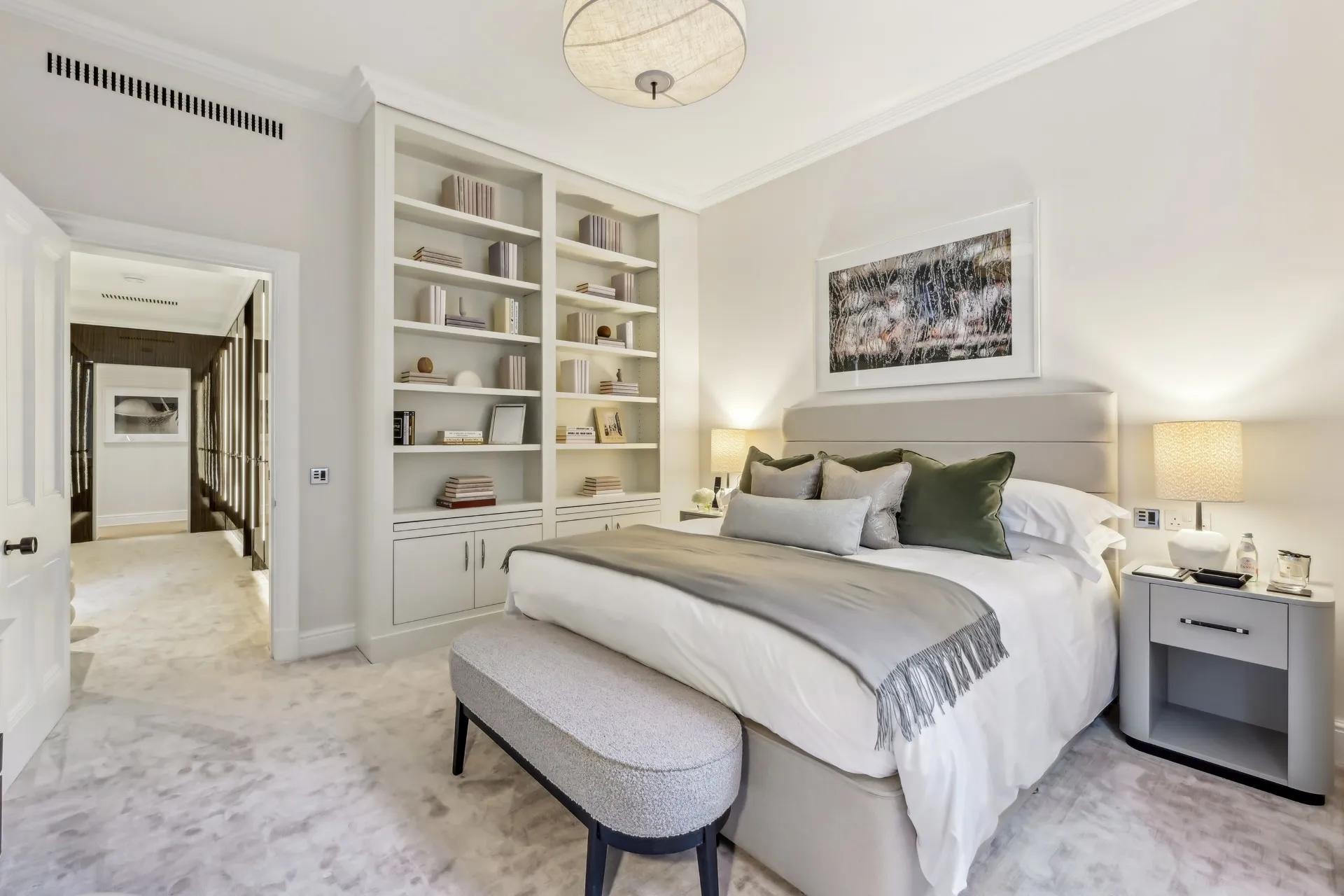 Calm bedroom with upholstered bed, built-in bookshelves, bedside lamps, and a soft bench.