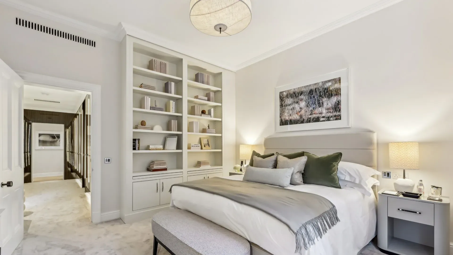 Calm bedroom with upholstered bed, built-in bookshelves, bedside lamps, and a soft bench.