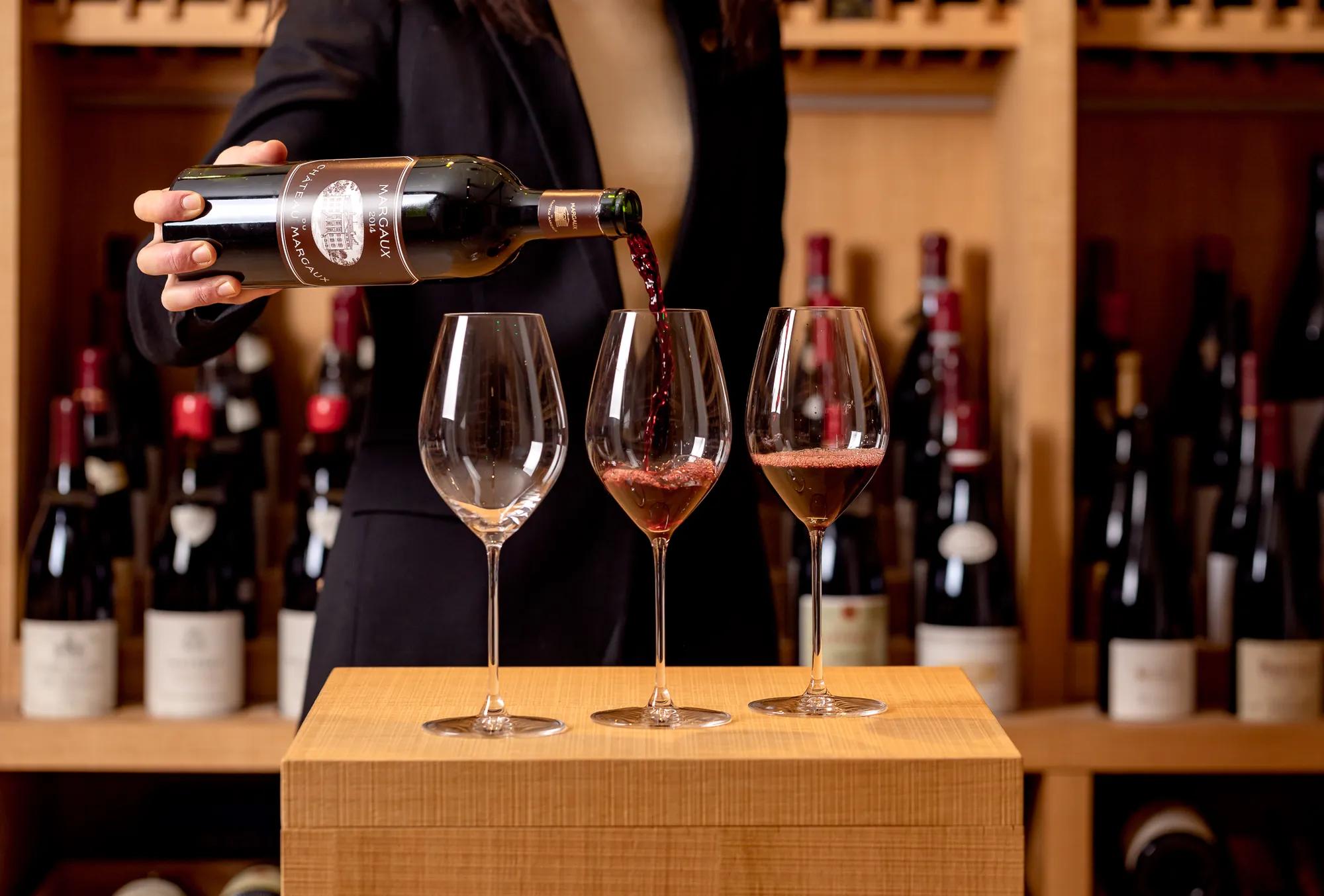 Person pouring red wine into three glasses in a wine cellar with bottles displayed behind.