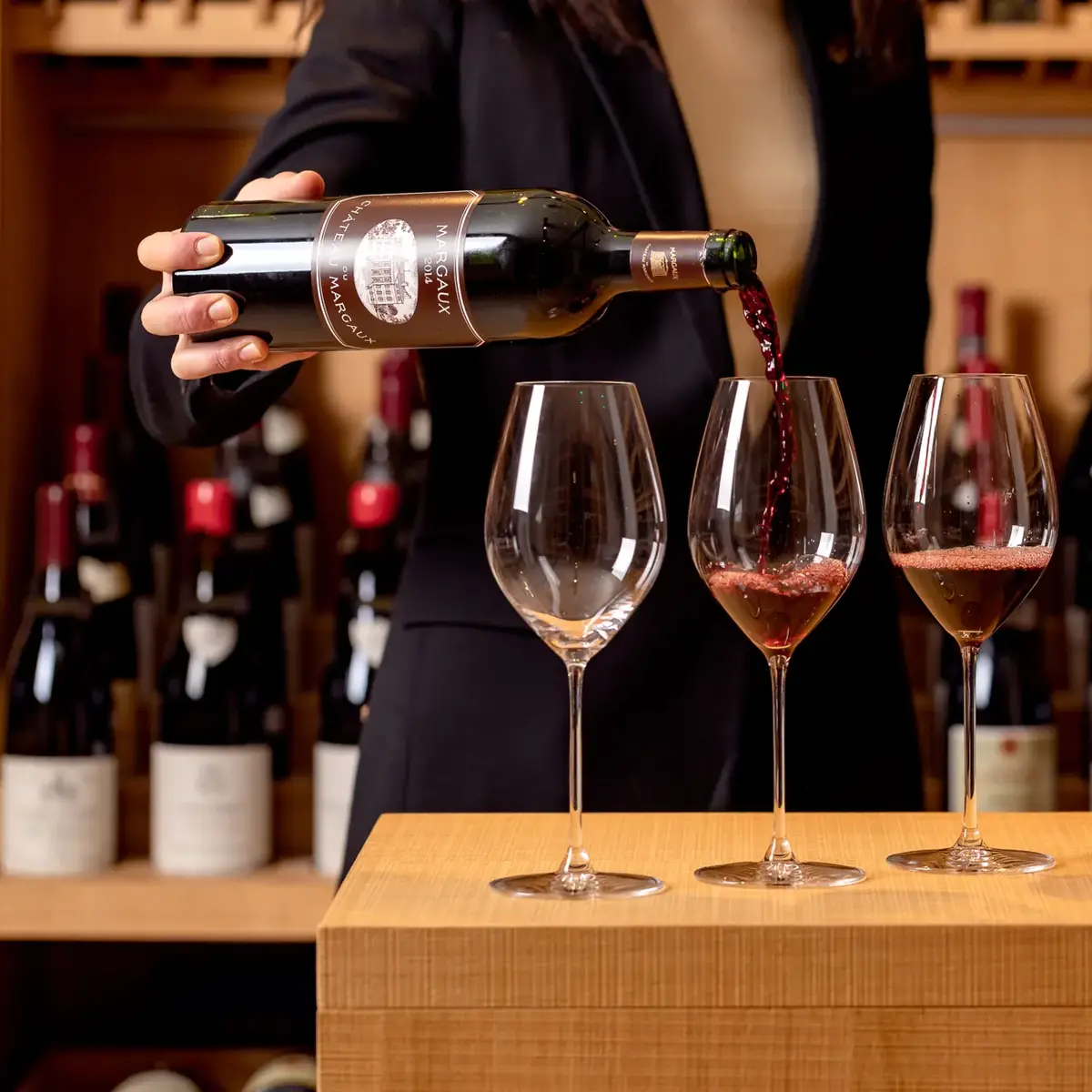 Person pouring red wine into three glasses in a wine cellar with bottles displayed behind.