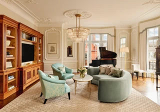 Elegant living room with a curved mint-green sofa, two teal armchairs, and a grand piano by tall windows. The room features ornate wall panelling, a crystal chandelier, and built-in wooden cabinetry with books and a television.
