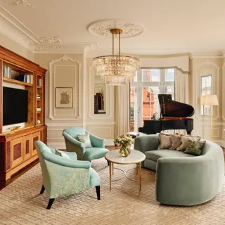 Elegant living room with a curved mint-green sofa, two teal armchairs, and a grand piano by tall windows. The room features ornate wall panelling, a crystal chandelier, and built-in wooden cabinetry with books and a television.