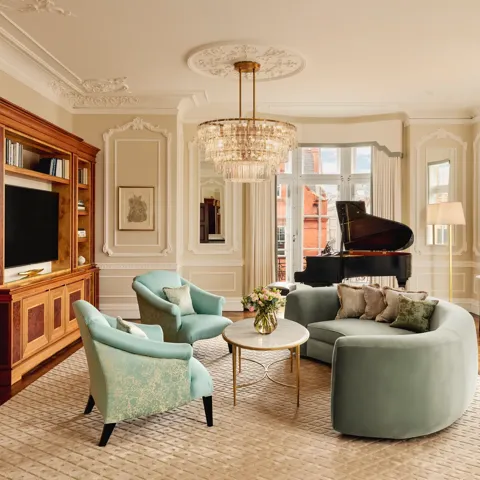 Elegant living room with a curved mint-green sofa, two teal armchairs, and a grand piano by tall windows. The room features ornate wall panelling, a crystal chandelier, and built-in wooden cabinetry with books and a television.