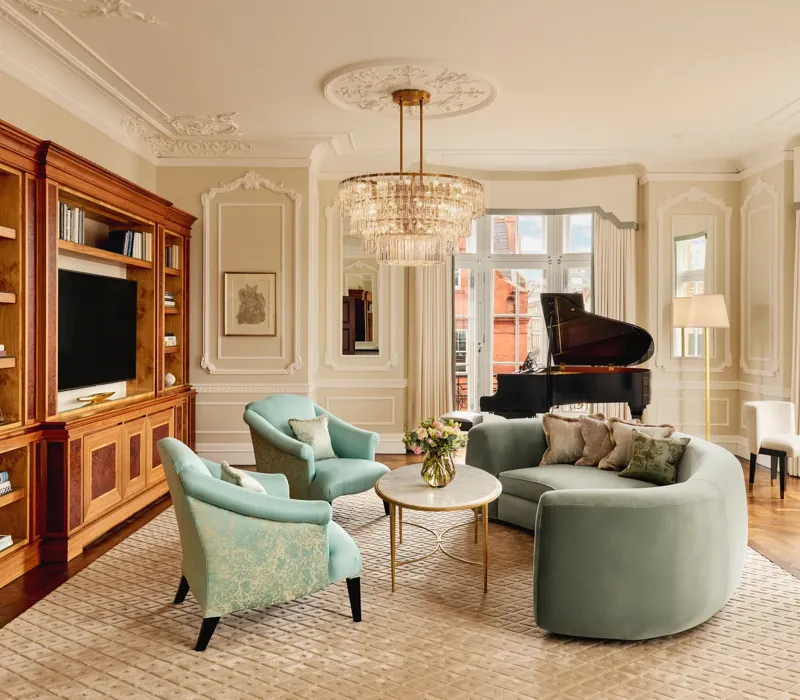 Elegant living room with a curved mint-green sofa, two teal armchairs, and a grand piano by tall windows. The room features ornate wall panelling, a crystal chandelier, and built-in wooden cabinetry with books and a television.
