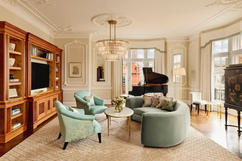 Elegant living room with a curved mint-green sofa, two teal armchairs, and a grand piano by tall windows. The room features ornate wall panelling, a crystal chandelier, and built-in wooden cabinetry with books and a television.