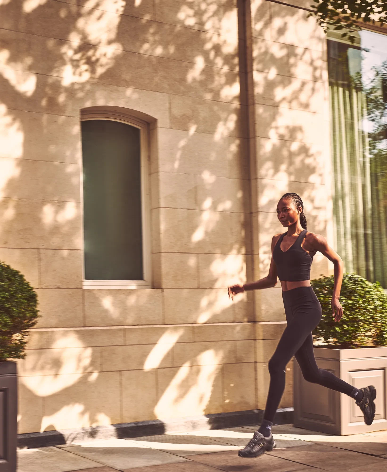 Woman in black athletic wear jogging along sun-dappled stone building exterior.