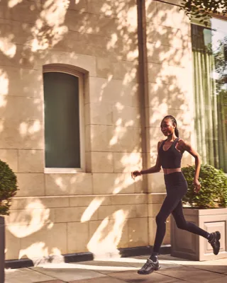Woman in black athletic wear jogging along sun-dappled stone building exterior.