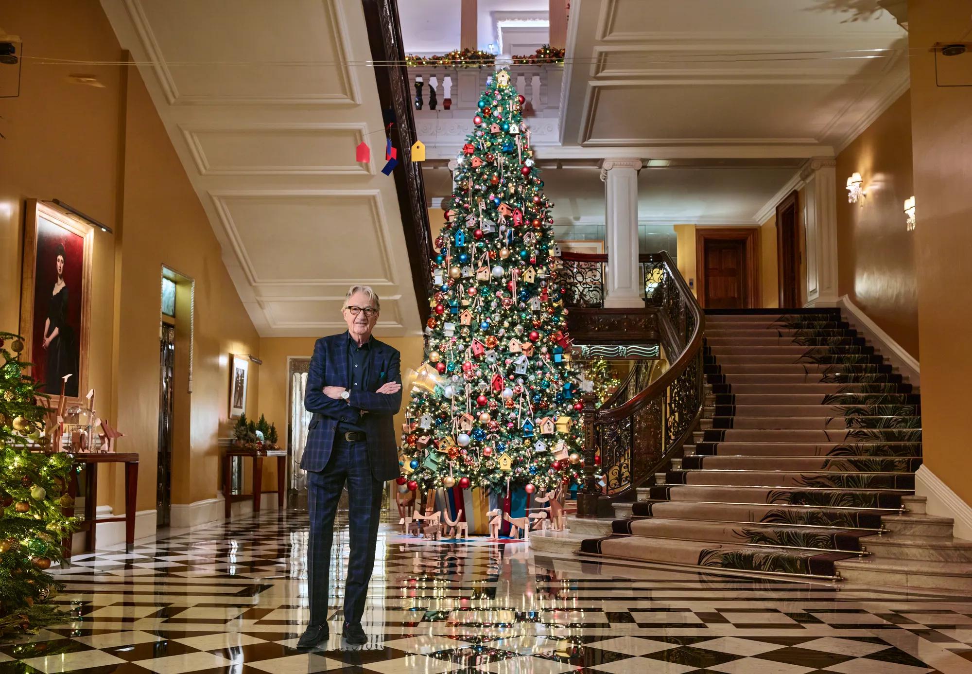 Grand hotel lobby with sweeping staircase and checkered floor, featuring a tall decorated Christmas tree and a man standing in foreground