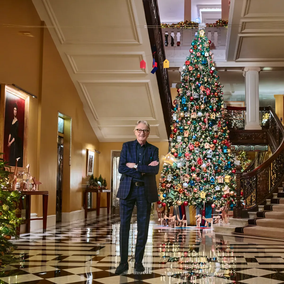 Grand hotel lobby with sweeping staircase and checkered floor, featuring a tall decorated Christmas tree and a man standing in foreground