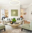 Bright contemporary living room featuring a curved cream sofa with silver cushions, two pale green armchairs, and a round coffee table topped with books, candles, and a green floral arrangement, set beneath a modern circular light fixture and minimalist wall art.