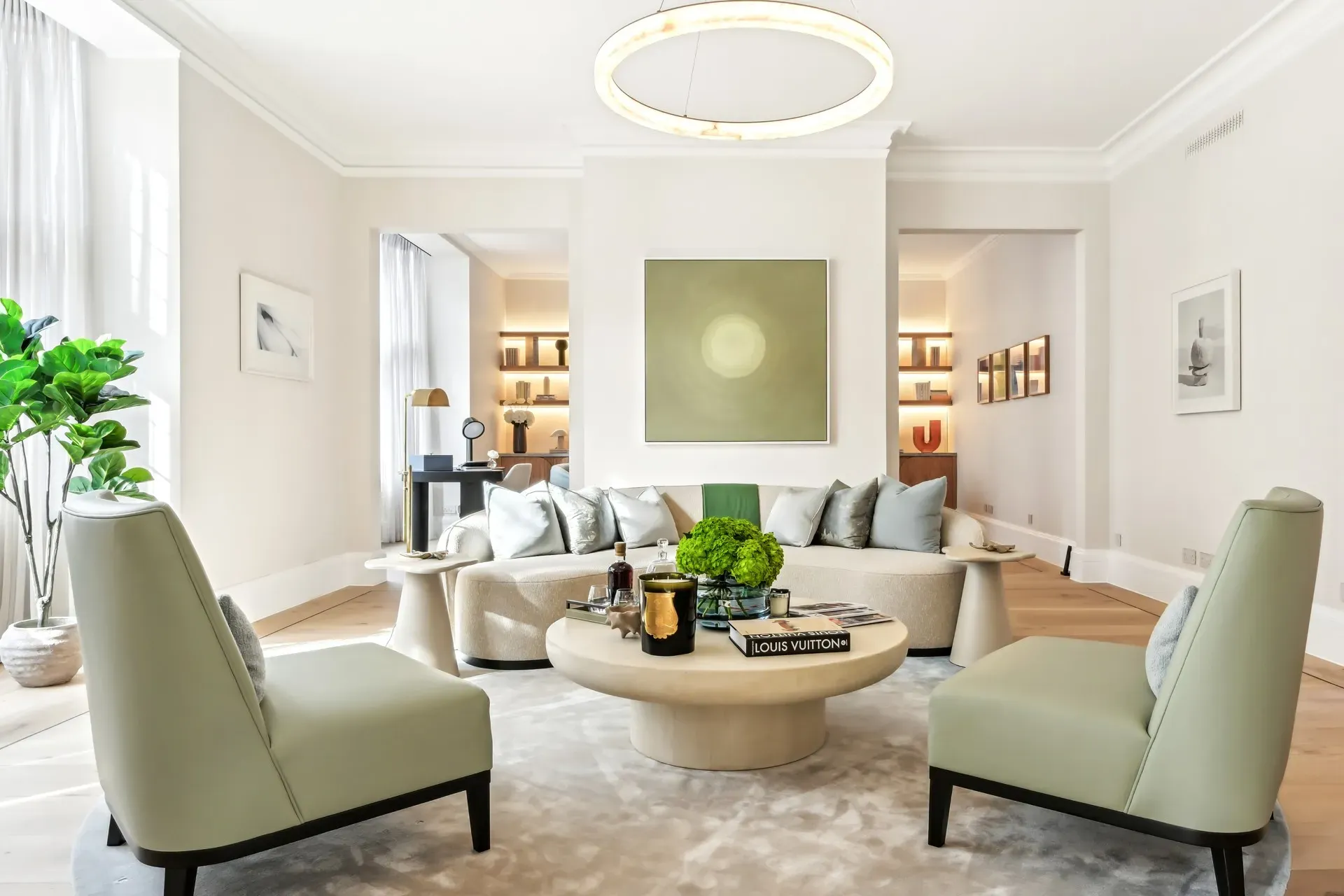 Bright contemporary living room featuring a curved cream sofa with silver cushions, two pale green armchairs, and a round coffee table topped with books, candles, and a green floral arrangement, set beneath a modern circular light fixture and minimalist wall art.