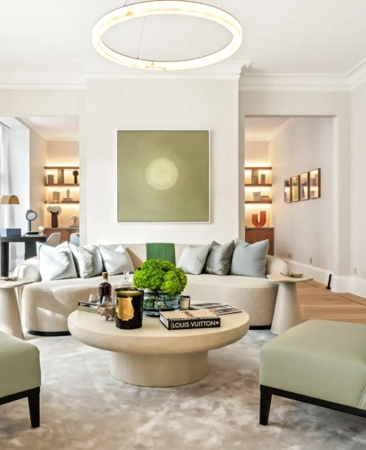 Bright contemporary living room featuring a curved cream sofa with silver cushions, two pale green armchairs, and a round coffee table topped with books, candles, and a green floral arrangement, set beneath a modern circular light fixture and minimalist wall art.