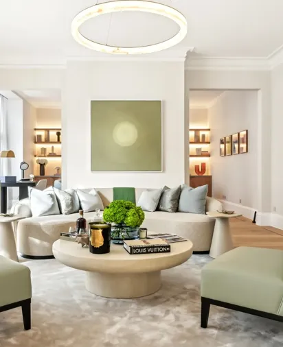 Bright contemporary living room featuring a curved cream sofa with silver cushions, two pale green armchairs, and a round coffee table topped with books, candles, and a green floral arrangement, set beneath a modern circular light fixture and minimalist wall art.
