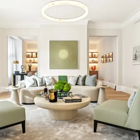 Bright contemporary living room featuring a curved cream sofa with silver cushions, two pale green armchairs, and a round coffee table topped with books, candles, and a green floral arrangement, set beneath a modern circular light fixture and minimalist wall art.