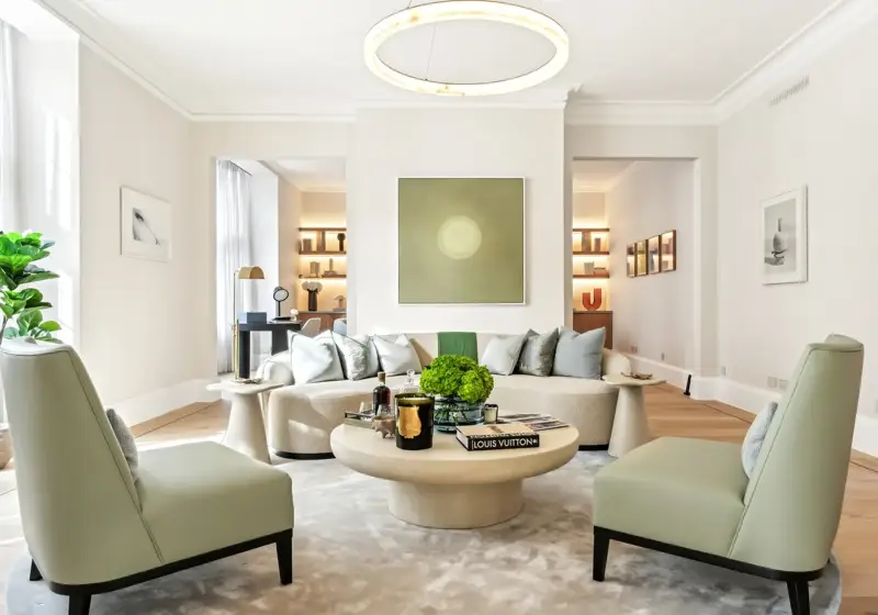 Bright contemporary living room featuring a curved cream sofa with silver cushions, two pale green armchairs, and a round coffee table topped with books, candles, and a green floral arrangement, set beneath a modern circular light fixture and minimalist wall art.
