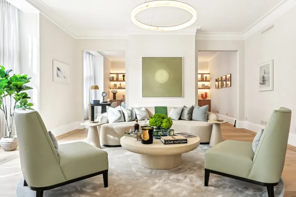 Bright contemporary living room featuring a curved cream sofa with silver cushions, two pale green armchairs, and a round coffee table topped with books, candles, and a green floral arrangement, set beneath a modern circular light fixture and minimalist wall art.