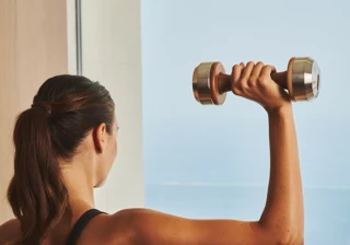 Person lifting a dumbbell indoors, facing a window with a sea view.