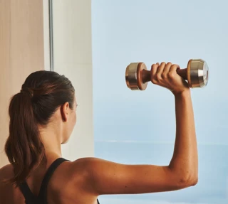 Person lifting a dumbbell indoors, facing a window with a sea view.