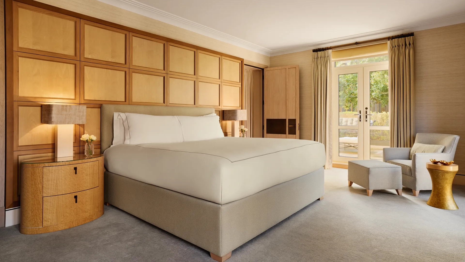 Spacious bedroom with a large bed framed by wooden paneling, a reading chair with ottoman, and French doors opening to a private terrace.