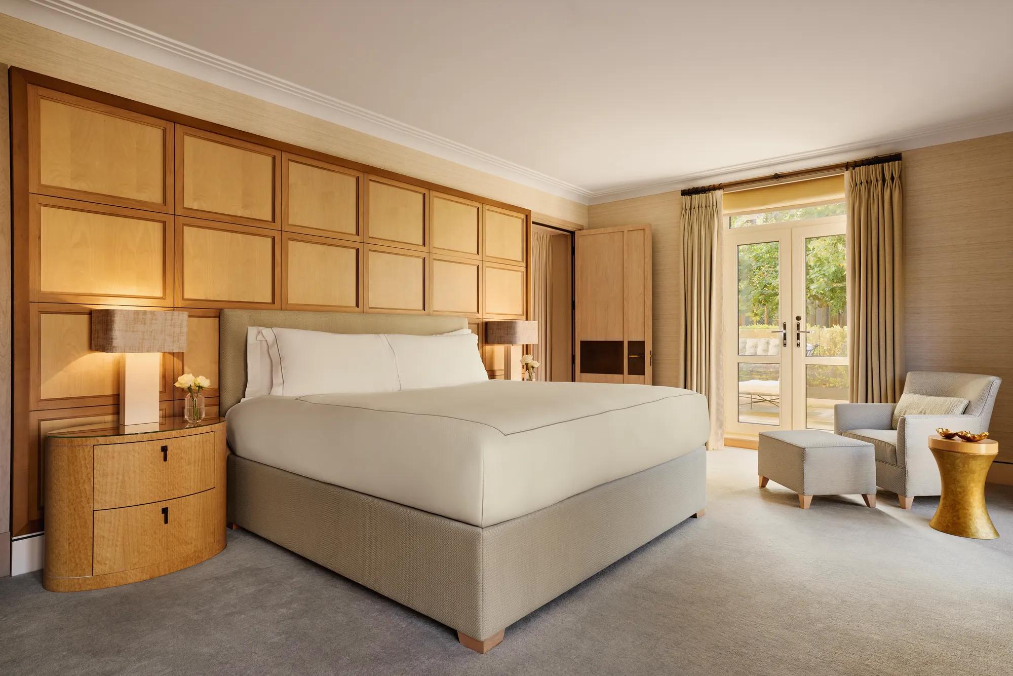 Spacious bedroom with a large bed framed by wooden paneling, a reading chair with ottoman, and French doors opening to a private terrace.