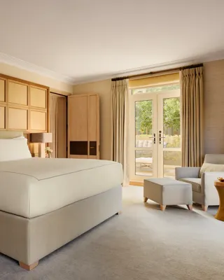Spacious bedroom with a large bed framed by wooden paneling, a reading chair with ottoman, and French doors opening to a private terrace.