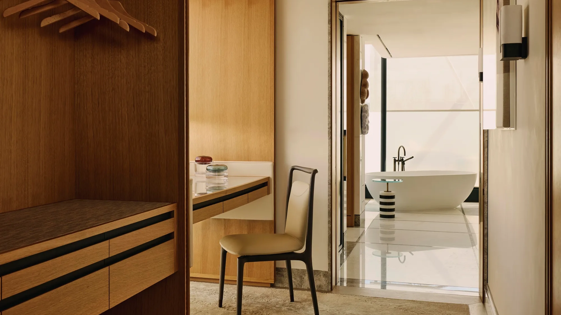 A dressing area with open wardrobes and a desk leading to a bright bathroom featuring a freestanding bathtub and modern fixtures.