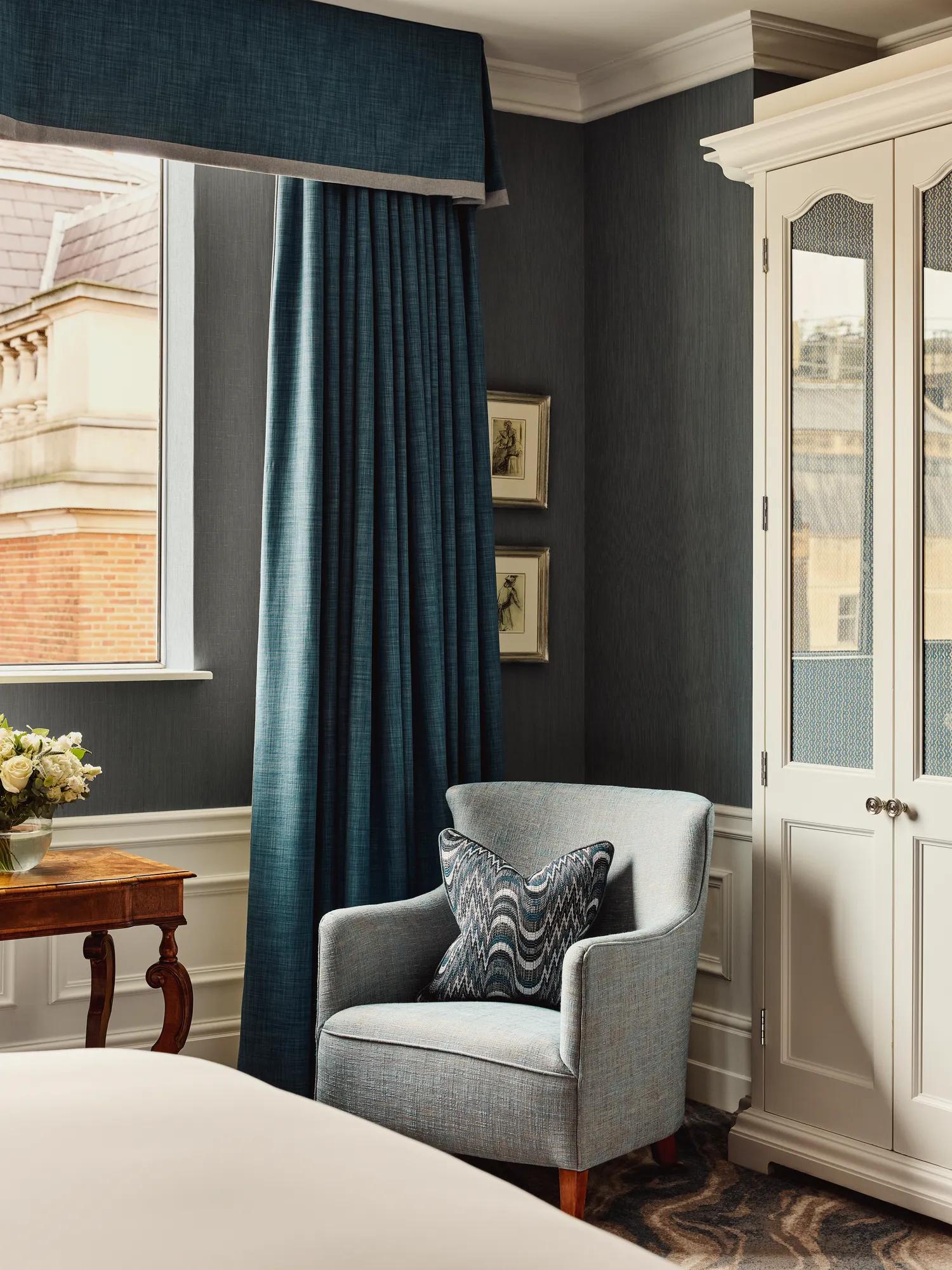 Bedroom corner with upholstered armchair, patterned cushion, blue curtains, window view, and white wardrobe in a classic hotel interior.