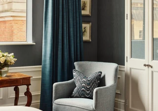 Bedroom corner with upholstered armchair, patterned cushion, blue curtains, window view, and white wardrobe in a classic hotel interior.