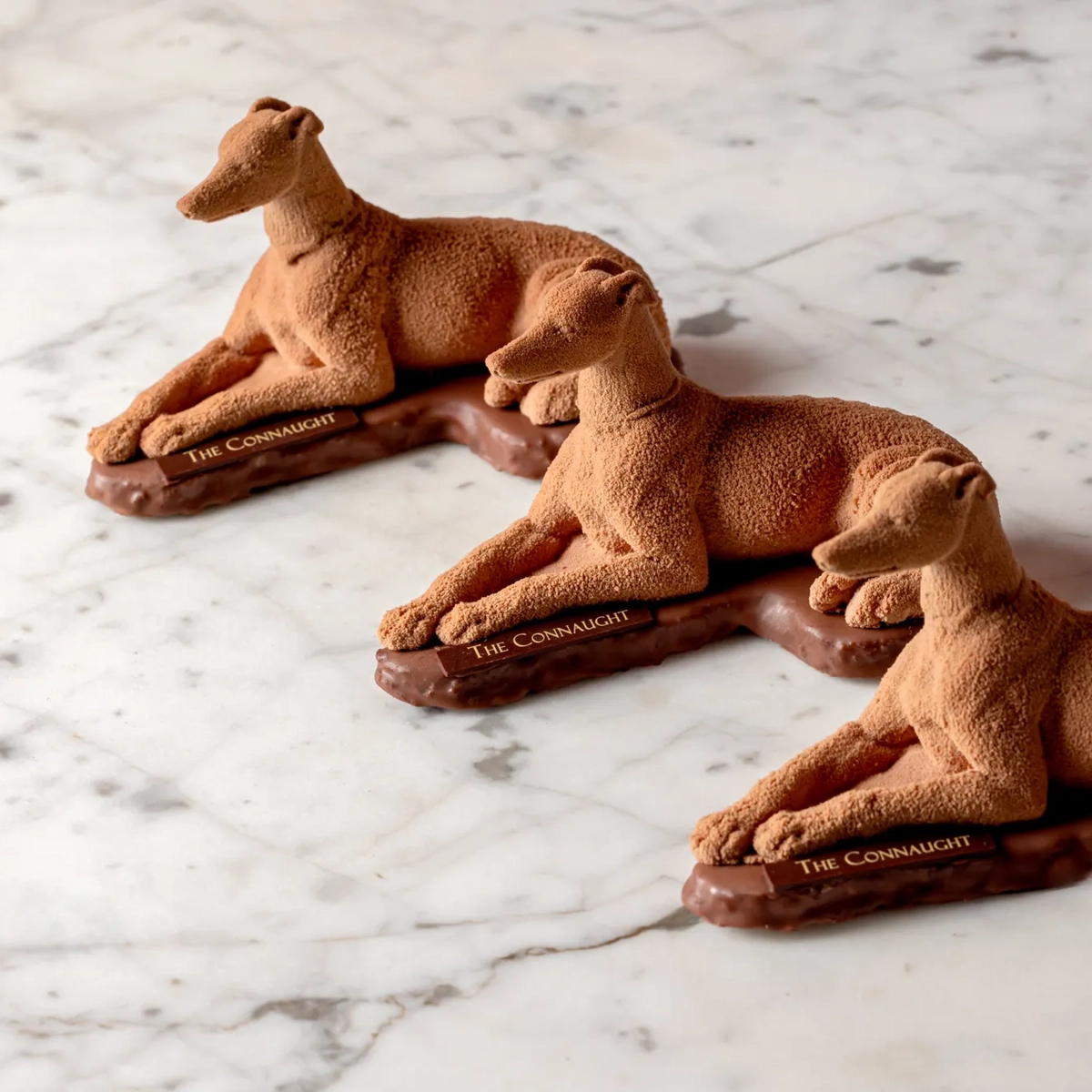 Three chocolate sculptures shaped like reclining greyhound dogs displayed on a marble surface.