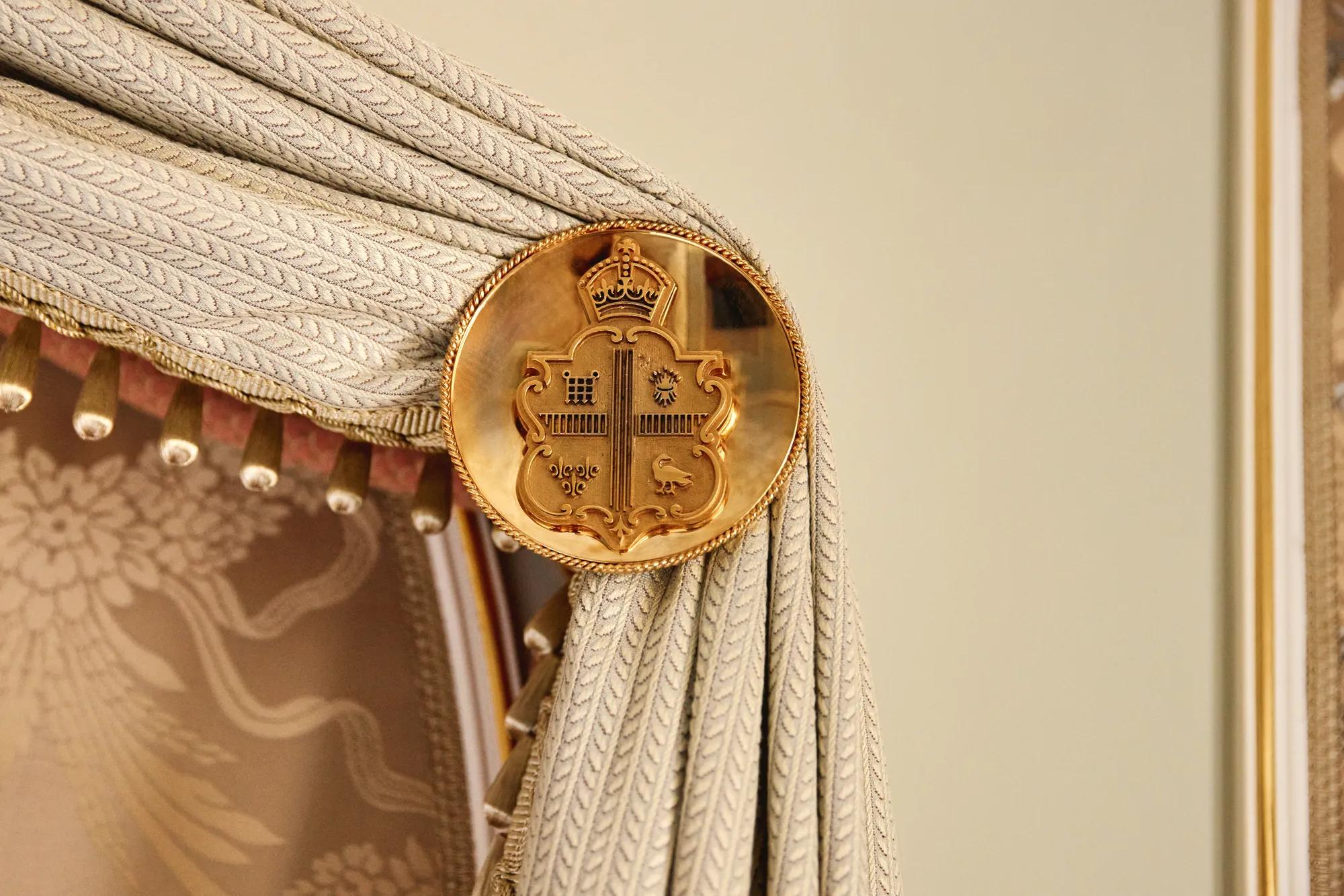 Close-up of an ornate gold medallion embossed with a crest, used as a curtain tieback holding pleated fabric with a textured braid pattern and tassel trim.