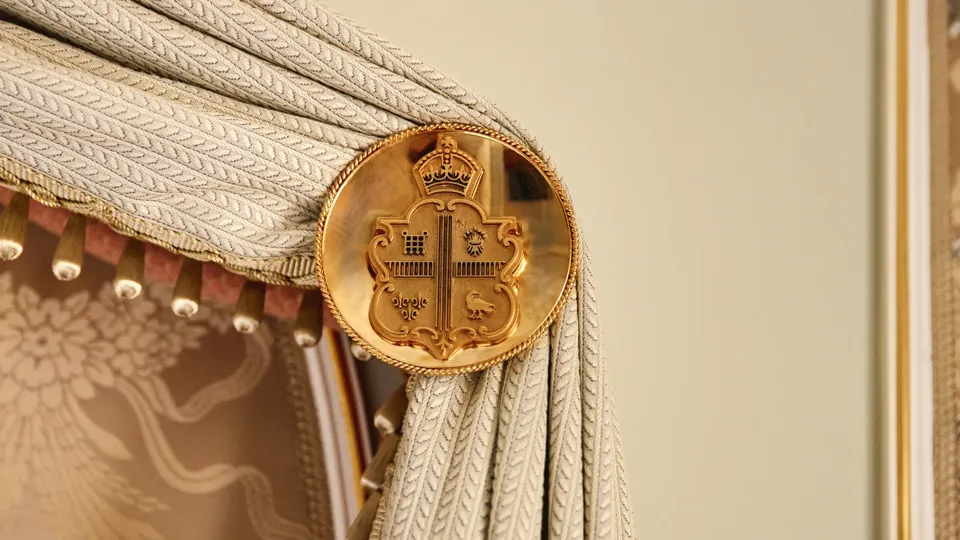 Close-up of an ornate gold medallion embossed with a crest, used as a curtain tieback holding pleated fabric with a textured braid pattern and tassel trim.