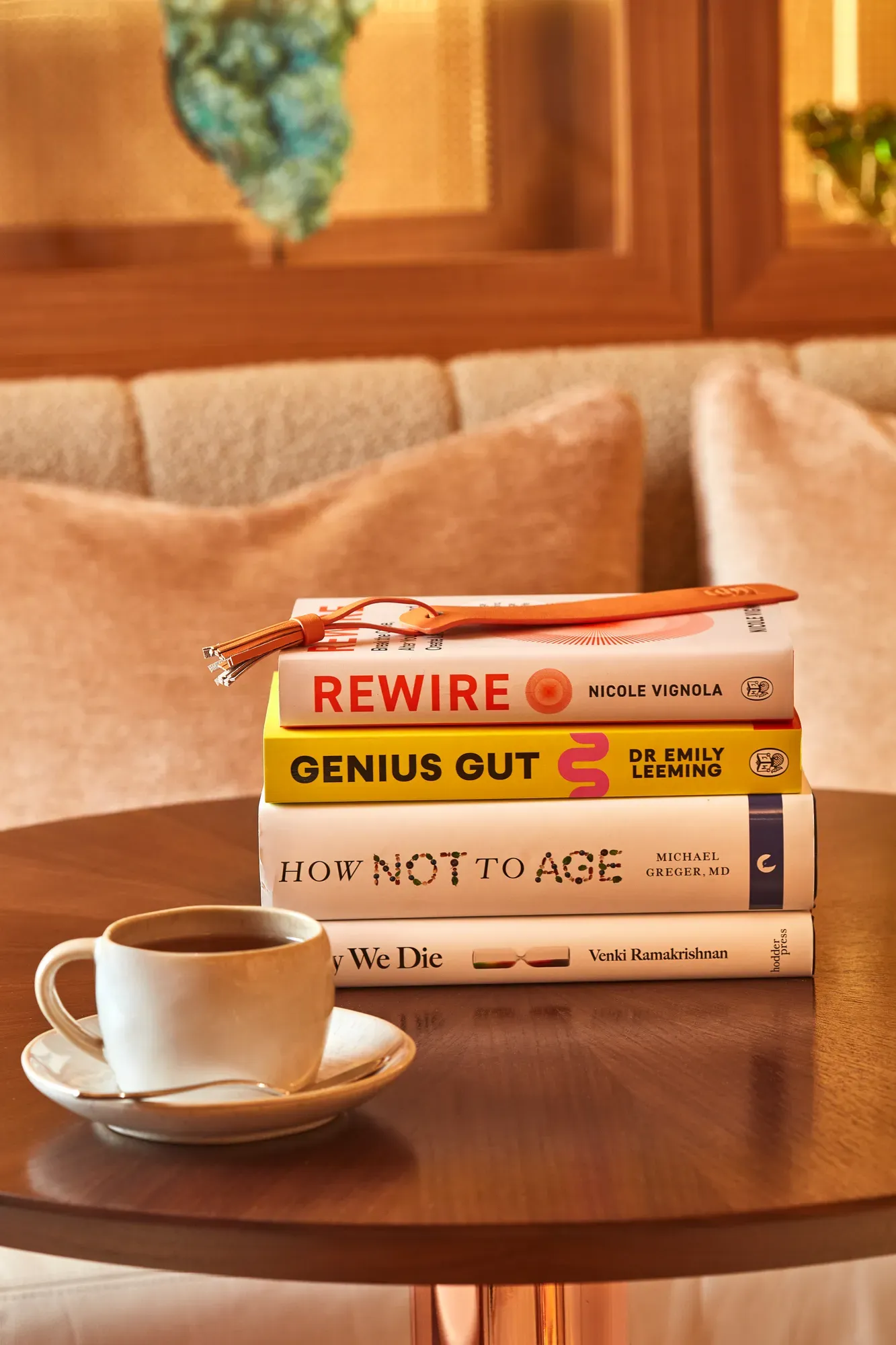 Stack of books with glasses on top beside a coffee cup on a round wooden table in a softly lit lounge.