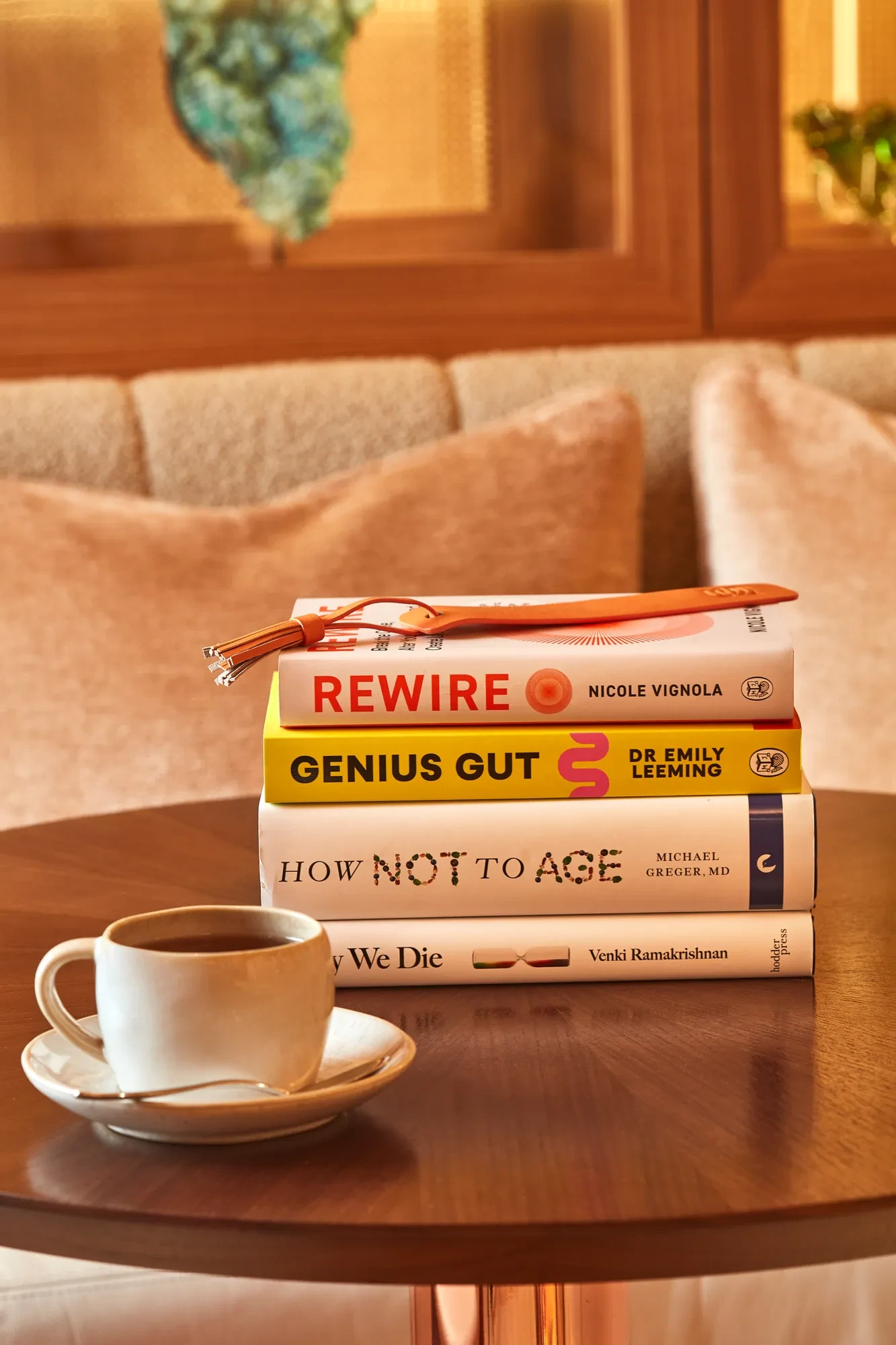 Stack of books with glasses on top beside a coffee cup on a round wooden table in a softly lit lounge.