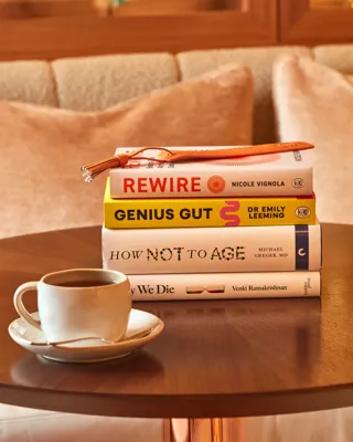 Stack of books with glasses on top beside a coffee cup on a round wooden table in a softly lit lounge.