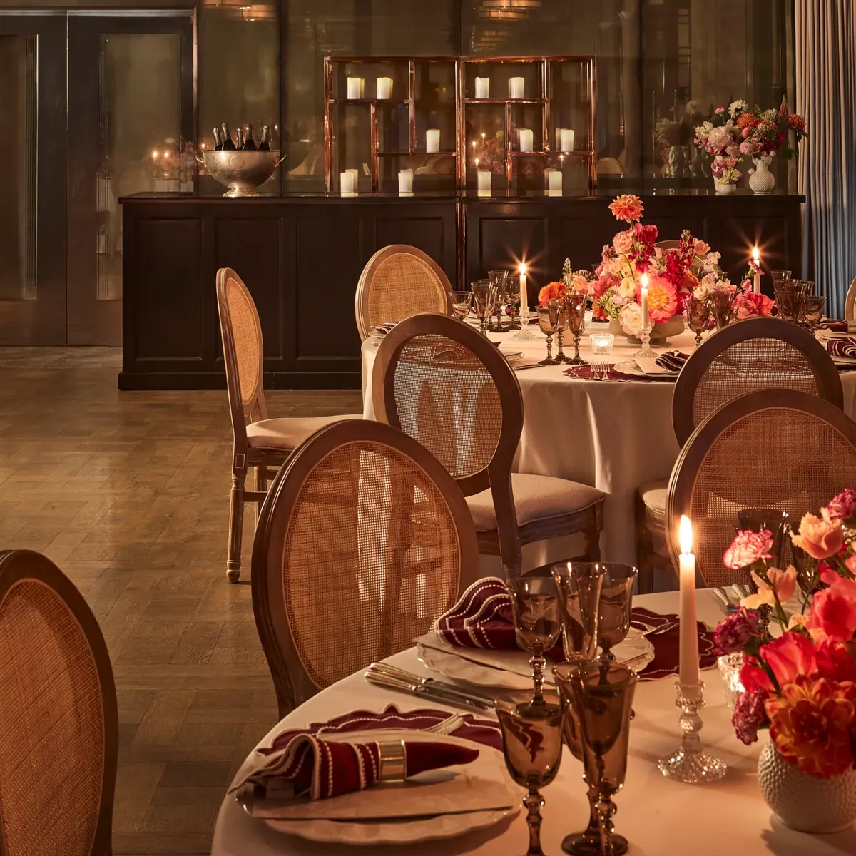 Elegant dining room with round tables set with flowers and candles, surrounded by cane-back chairs under warm ambient lighting.