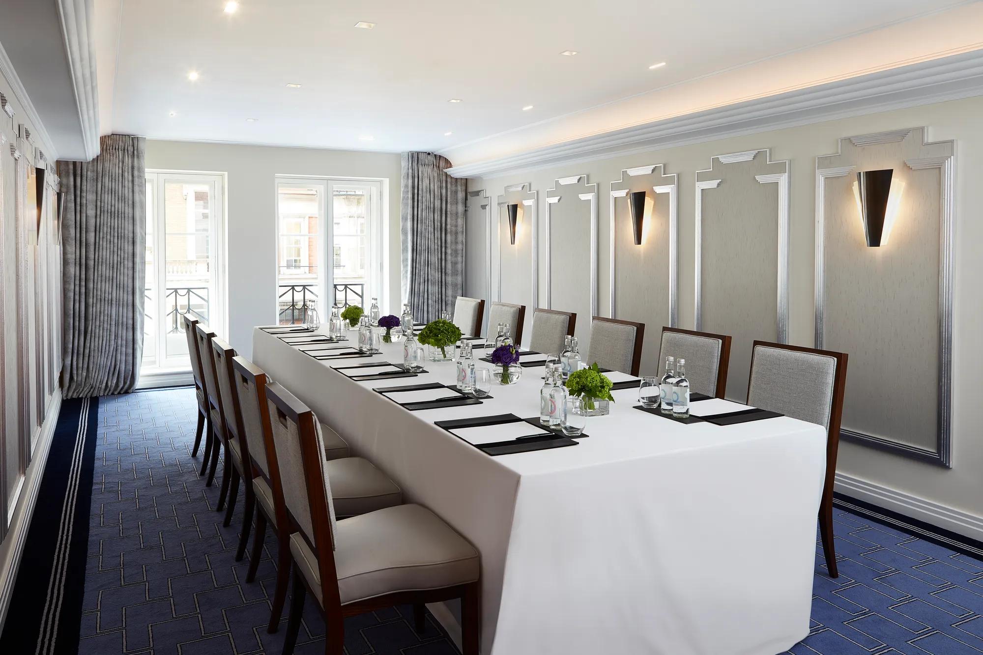 Bright meeting room with a long U-shaped table covered in white linen, surrounded by upholstered chairs, and set with notepads, glass bottles, and small green and purple floral arrangements beneath modern wall sconces.