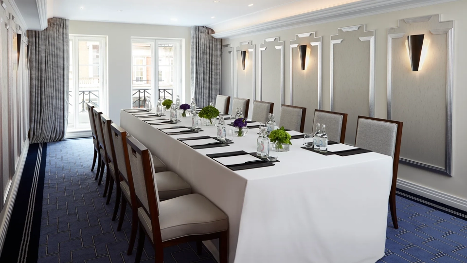 Bright meeting room with a long U-shaped table covered in white linen, surrounded by upholstered chairs, and set with notepads, glass bottles, and small green and purple floral arrangements beneath modern wall sconces.