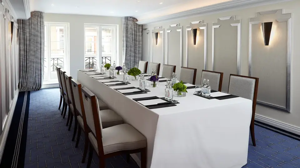Bright meeting room with a long U-shaped table covered in white linen, surrounded by upholstered chairs, and set with notepads, glass bottles, and small green and purple floral arrangements beneath modern wall sconces.