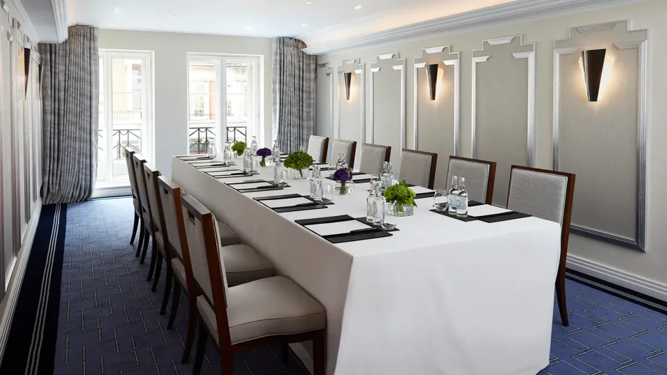 Bright meeting room with a long U-shaped table covered in white linen, surrounded by upholstered chairs, and set with notepads, glass bottles, and small green and purple floral arrangements beneath modern wall sconces.