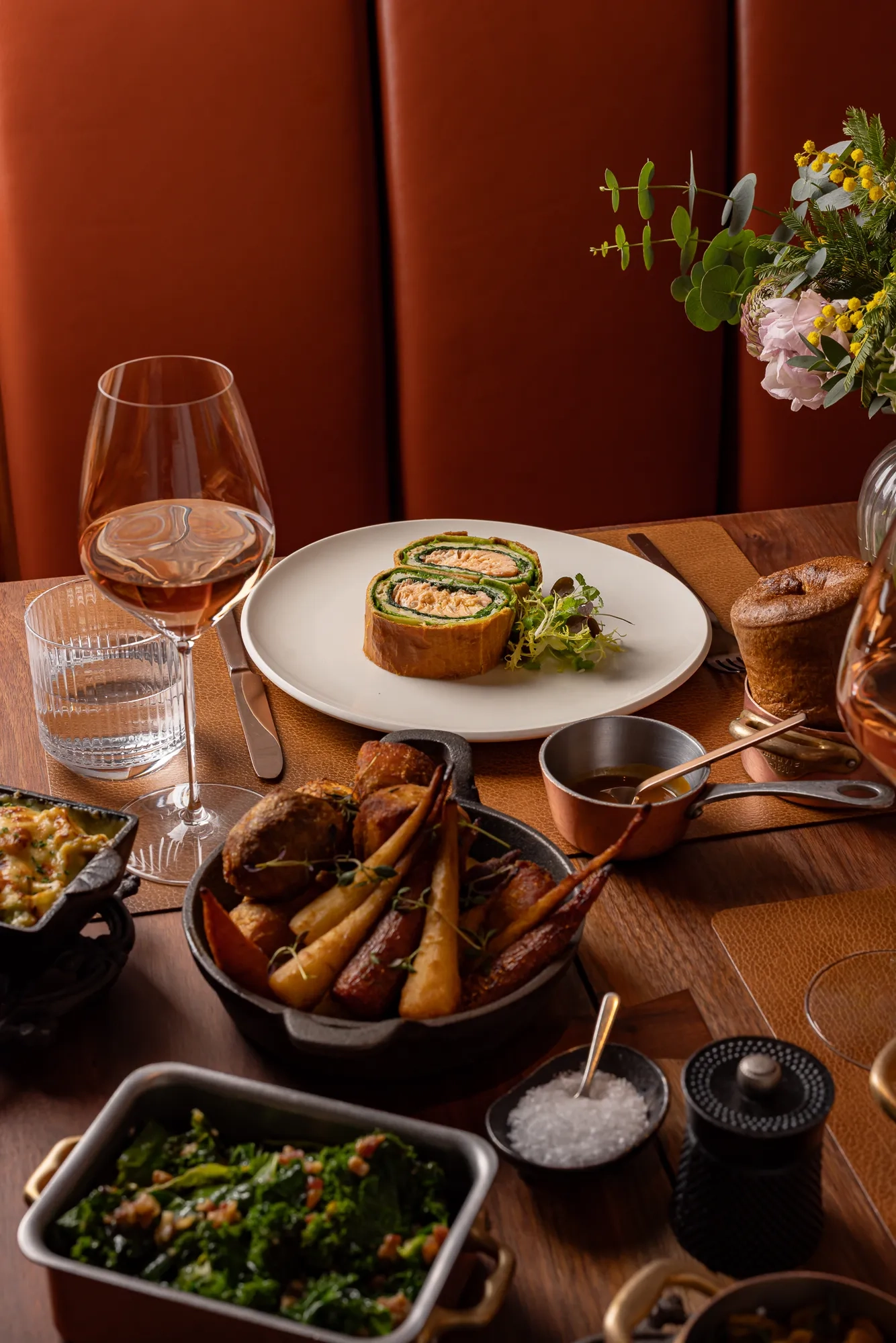 Salmon Wellington served with greens, roast vegetables, sides, and wine on a warmly lit wooden dining table.