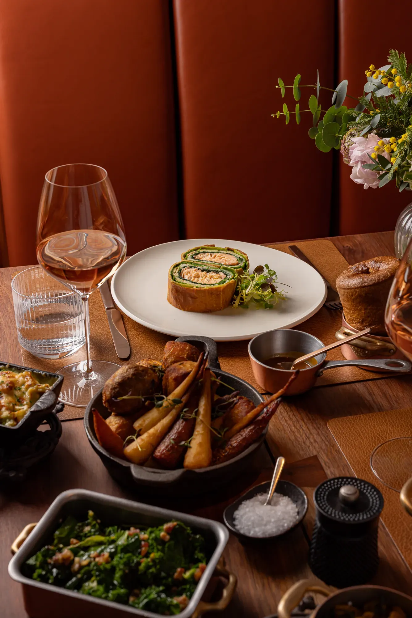Salmon Wellington served with greens, roast vegetables, sides, and wine on a warmly lit wooden dining table.
