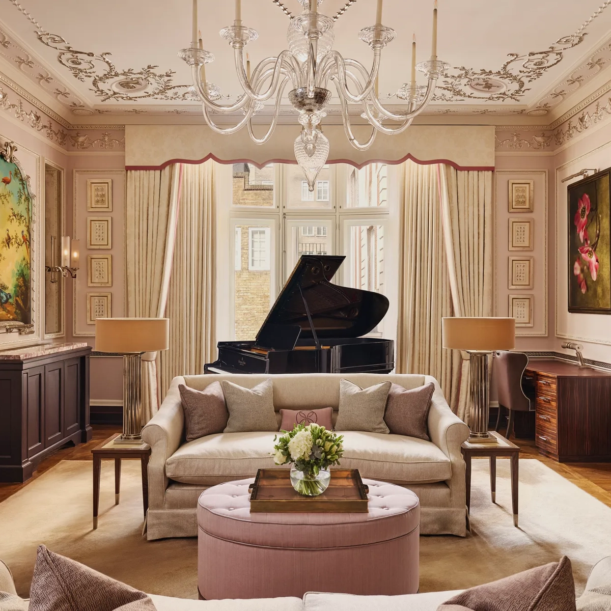 An elegant living room with blush-toned walls, ornate ceiling mouldings, and a grand piano by the window, featuring cream sofas, a pink ottoman, and crystal chandelier under soft natural light.