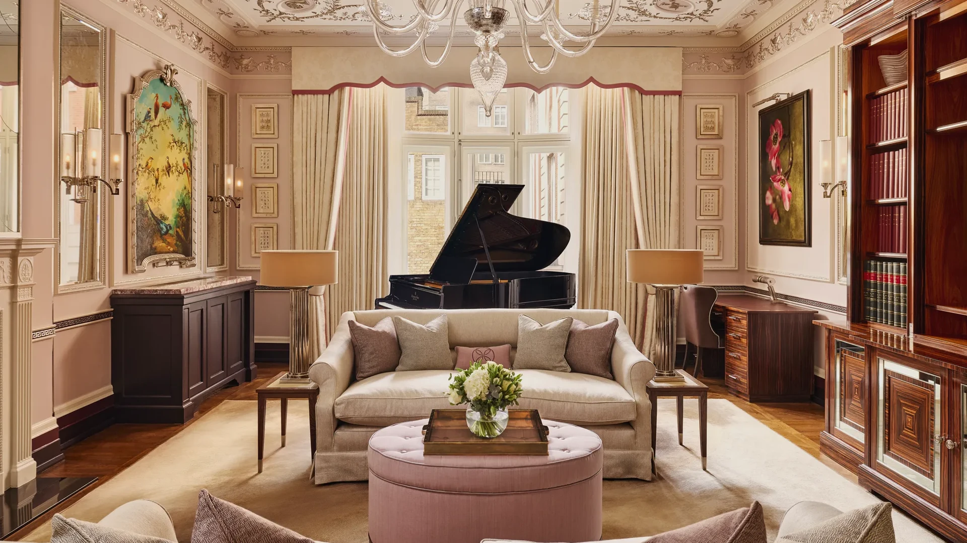 An elegant living room with blush-toned walls, ornate ceiling mouldings, and a grand piano by the window, featuring cream sofas, a pink ottoman, and crystal chandelier under soft natural light.