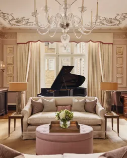 An elegant living room with blush-toned walls, ornate ceiling mouldings, and a grand piano by the window, featuring cream sofas, a pink ottoman, and crystal chandelier under soft natural light.
