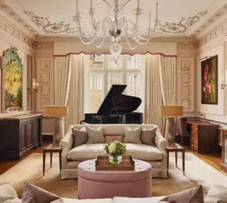 An elegant living room with blush-toned walls, ornate ceiling mouldings, and a grand piano by the window, featuring cream sofas, a pink ottoman, and crystal chandelier under soft natural light.