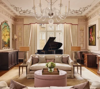 An elegant living room with blush-toned walls, ornate ceiling mouldings, and a grand piano by the window, featuring cream sofas, a pink ottoman, and crystal chandelier under soft natural light.
