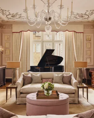 An elegant living room with blush-toned walls, ornate ceiling mouldings, and a grand piano by the window, featuring cream sofas, a pink ottoman, and crystal chandelier under soft natural light.