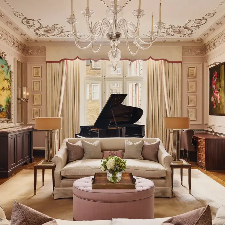 An elegant living room with blush-toned walls, ornate ceiling mouldings, and a grand piano by the window, featuring cream sofas, a pink ottoman, and crystal chandelier under soft natural light.