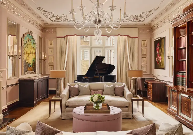 An elegant living room with blush-toned walls, ornate ceiling mouldings, and a grand piano by the window, featuring cream sofas, a pink ottoman, and crystal chandelier under soft natural light.