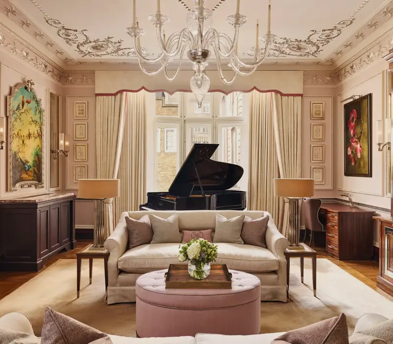 An elegant living room with blush-toned walls, ornate ceiling mouldings, and a grand piano by the window, featuring cream sofas, a pink ottoman, and crystal chandelier under soft natural light.